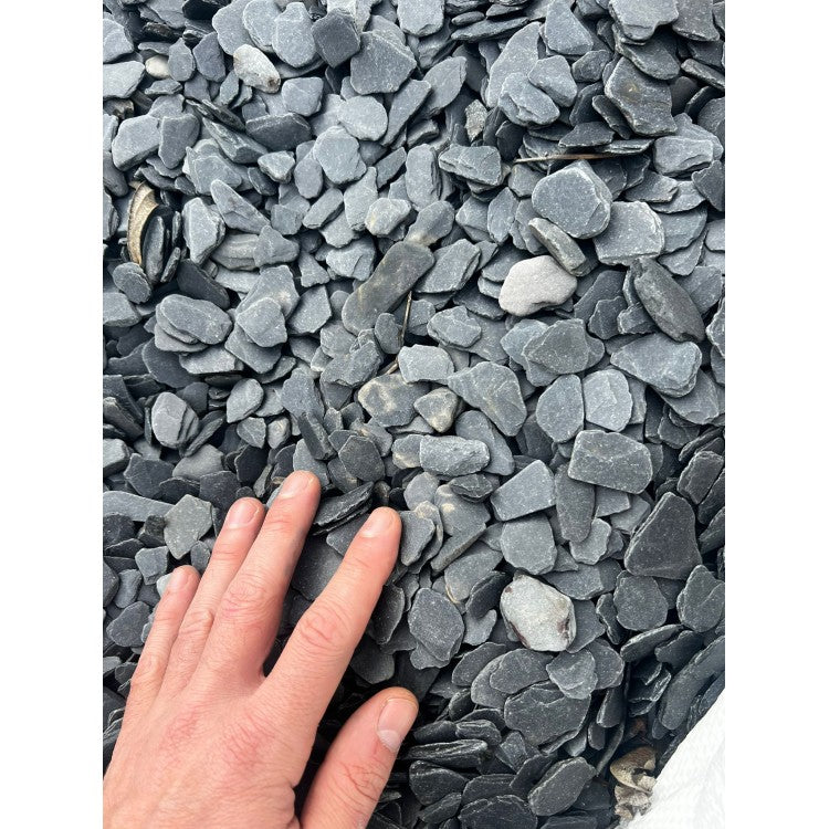 Das Bild zeigt eine Hand, die auf einer Ansammlung von Wild Rhino stones liegt. Die Steine sind unterschiedlich groß und haben eine unregelmäßige Form mit scharfen Kanten. Die Farbe der Schieferstücke ist ein dunkles Grau mit leicht bläulichen Schimmern. Die Oberfläche der Steine ist rau und uneben. Im Hintergrund ist ein weißer Sack zu erkennen, aus dem die Schieferstücke herausragen.